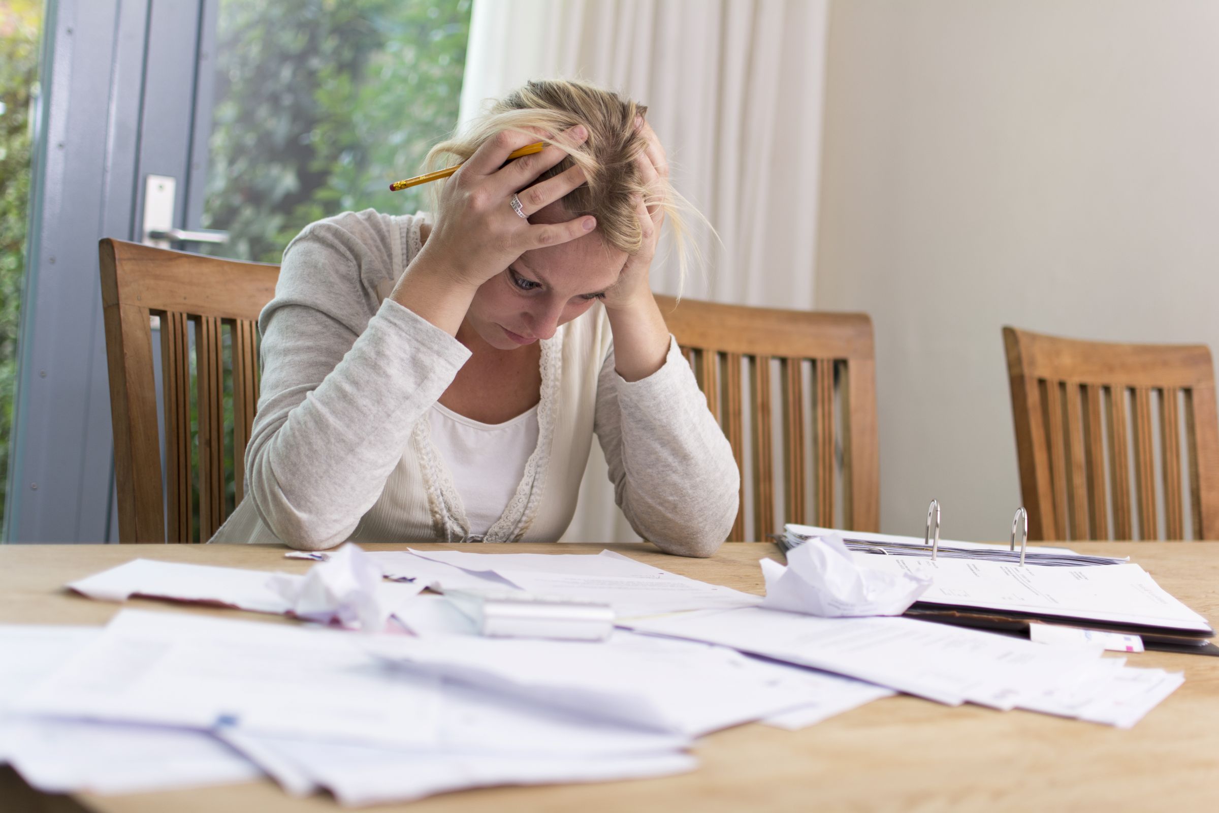 Woman sits at table holding her head in her hands in frustration as she looks at scattered bills.